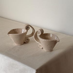 May include: Two ceramic cream pitchers with a speckled, light beige finish. Each pitcher has a spout and a decorative, scrolled handle. The pitchers are placed on a light-colored tablecloth.