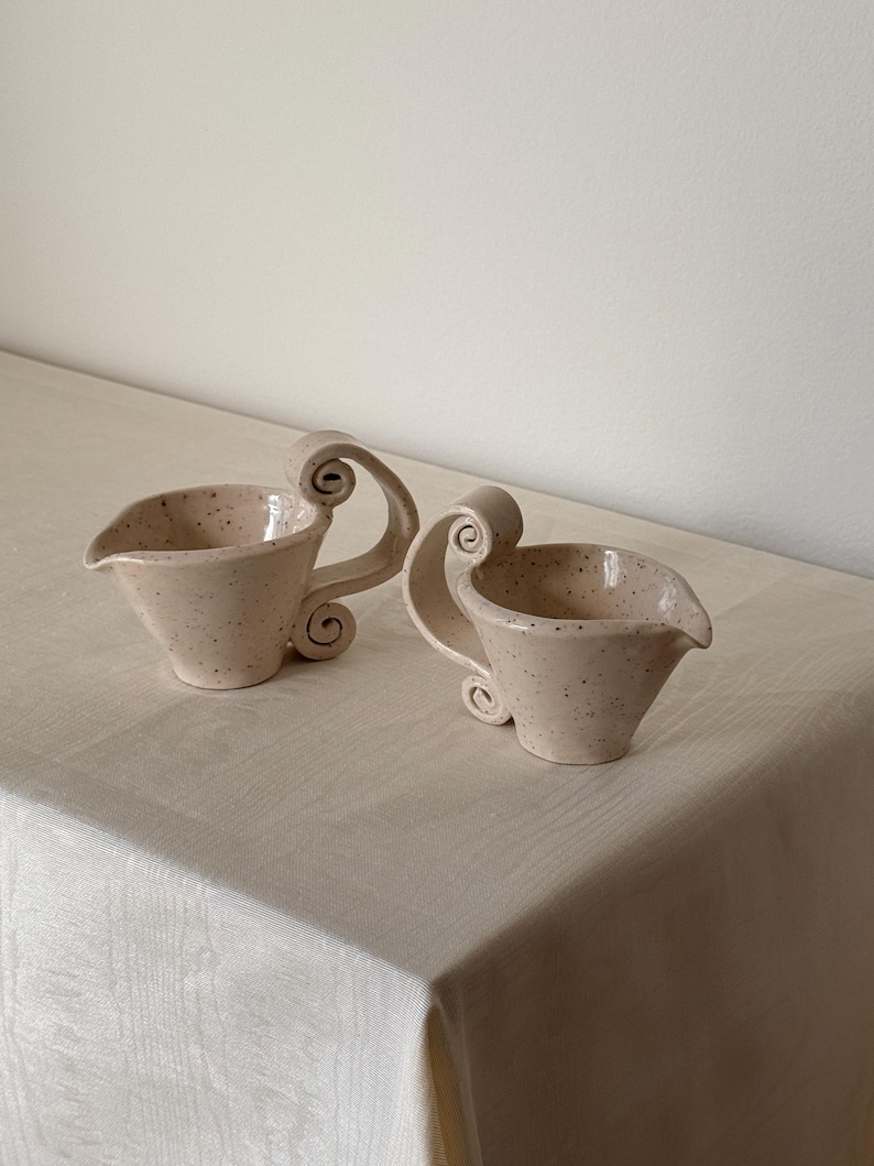 May include: Two ceramic cream pitchers with a speckled, light beige finish. Each pitcher has a spout and a decorative, scrolled handle. The pitchers are placed on a light-colored tablecloth.