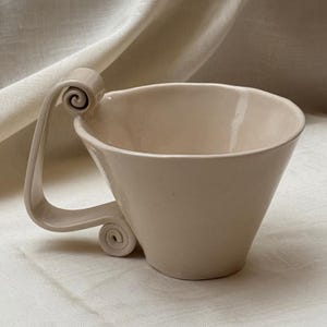 May include: A beige ceramic cup with a smooth, glossy finish and a wide, flared rim. The cup features a distinctive, large handle that extends upward and curves back down, ending in a decorative spiral scroll at the base of the vessel.