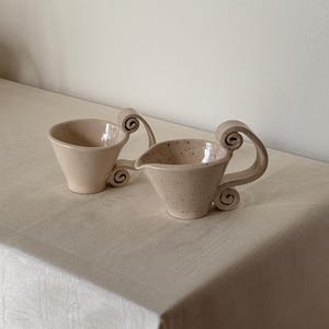 May include: A set of three ceramic mugs with a speckled, matte finish. The mugs are in shades of cream, light gray, and dark gray. Each mug has a simple, minimalist design and a rounded handle.