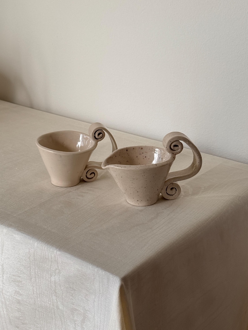 May include: A set of three ceramic mugs with a speckled, matte finish. The mugs are in shades of cream, light gray, and dark gray. Each mug has a simple, minimalist design and a rounded handle.