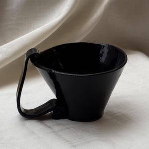 May include: A glossy black ceramic cup with a wide, flared rim and a distinctive, curved handle that loops from the top edge down to the base. The surface of the cup has a smooth, reflective finish, highlighting its deep, solid black color throughout.
