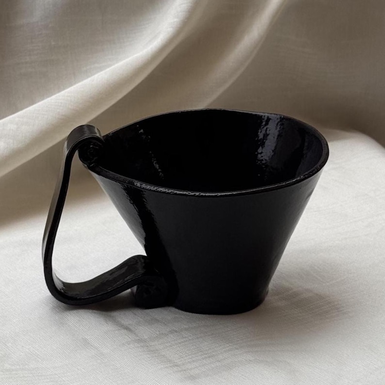 May include: A glossy black ceramic cup with a wide, flared rim and a distinctive, curved handle that loops from the top edge down to the base. The surface of the cup has a smooth, reflective finish, highlighting its deep, solid black color throughout.