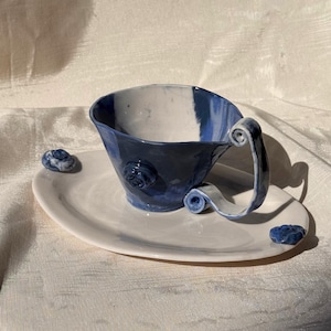 May include: A ceramic cup and saucer set featuring a marbled blue and white glaze. The cup has a flared rim, a curled handle, and a small rose-shaped appliqué on the side. The oval saucer includes two small blue rose-shaped accents on its surface.