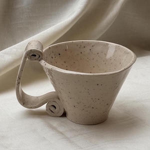 May include: A speckled cream-colored ceramic mug with a wide, flared rim and a unique, sculptural handle that features a prominent scroll design at the top and a smaller, tightly coiled spiral at the base, creating a distinct silhouette.