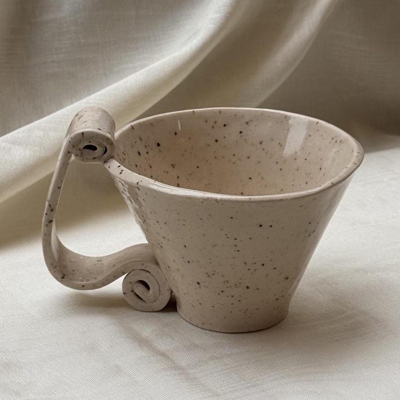 May include: A speckled cream-colored ceramic mug with a wide, flared rim and a unique, sculptural handle that features a prominent scroll design at the top and a smaller, tightly coiled spiral at the base, creating a distinct silhouette.