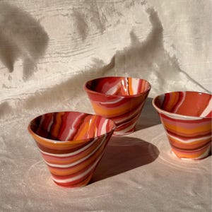 May include: Three ceramic cups featuring a swirling marbled pattern in shades of red, orange, pink, and white. Each cup has a wide, flared rim and a tapered base, showcasing a smooth, glossy finish with distinct, flowing horizontal color bands.