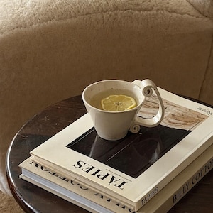 May include: A ceramic mug with a curled handle containing a light-colored liquid and a lemon slice, resting on two stacked hardcover books titled T&Agrave;PIES and BILLY COTTON. The books are placed on a dark brown circular wooden side table.
