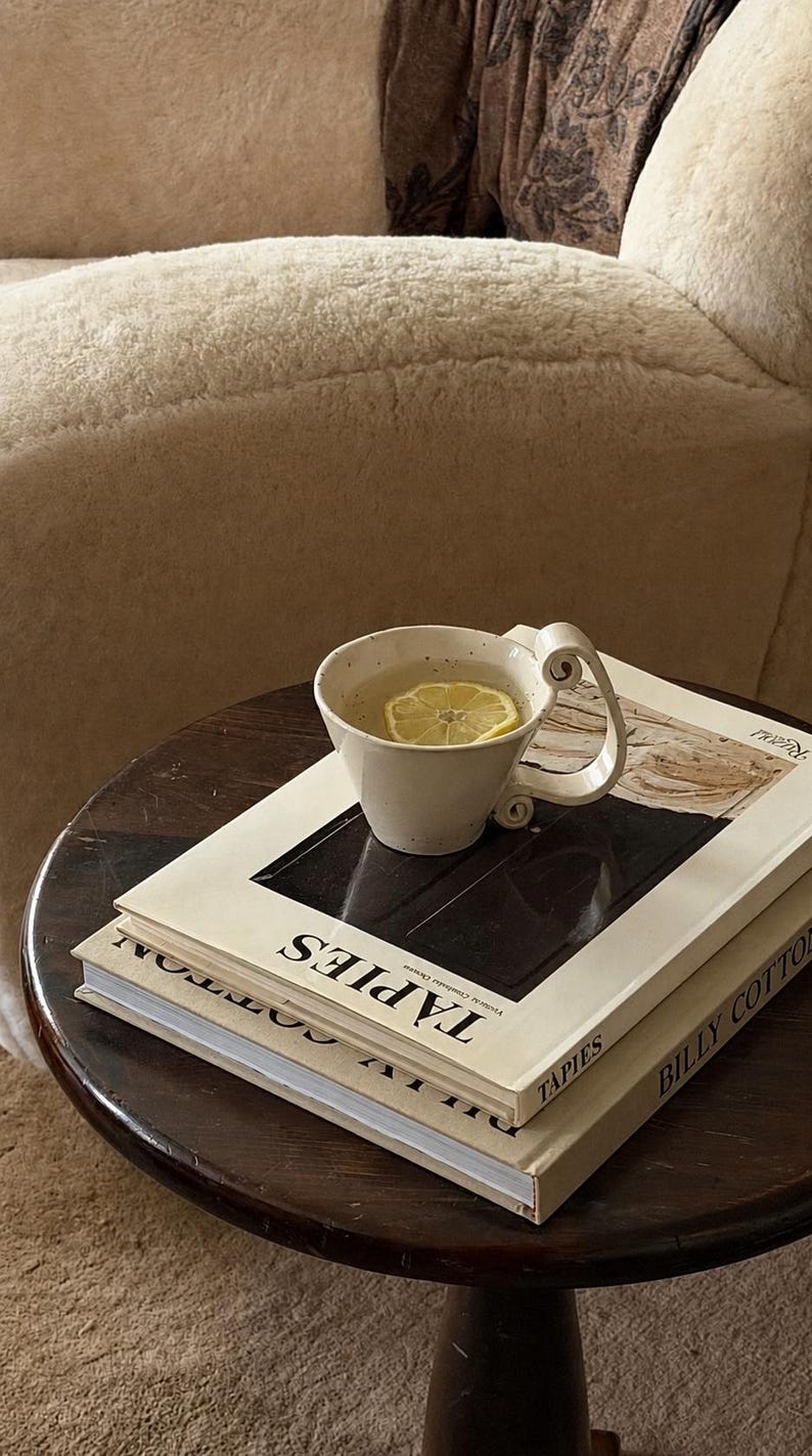 May include: A ceramic mug with a curled handle containing a light-colored liquid and a lemon slice, resting on two stacked hardcover books titled T&Agrave;PIES and BILLY COTTON. The books are placed on a dark brown circular wooden side table.