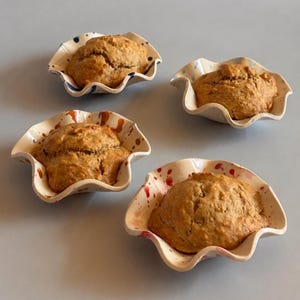 May include: Four ceramic bowls with ruffled edges, each containing a golden-brown baked muffin. The bowls feature distinct hand-painted patterns: blue dots, blue dashes, brown splatters, and red splatters on a cream-colored glazed surface.