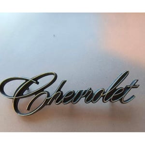 May include: A vintage Chevrolet emblem in a cursive script, with a dark metallic finish. The emblem is set against a neutral background, highlighting the classic design and automotive heritage.