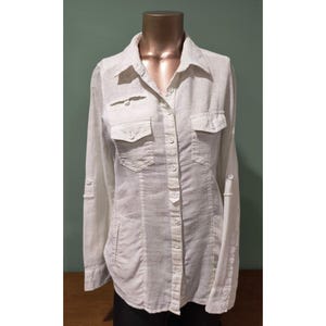 May include: A white linen button-down shirt with long sleeves, two chest pockets, and roll-up sleeve tabs. The shirt is displayed on a mannequin and has a classic collar and button-up front.