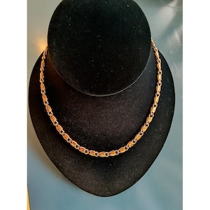 May include: A gold-colored necklace with an oval link design is displayed on a black velvet bust. The necklace is centered and hangs naturally, showcasing its intricate pattern and warm metallic hue. The background is a muted blue.