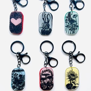 May include: Set of eight black metal keychains with colourful rectangular charms featuring various designs, including a heart, a rabbit, flowers, a dragonfly, a sunflower, a bird, and a landscape. The charms are labelled "Art + Aroma Diffuser Pendants".