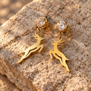 May include: A pair of gold-toned drop earrings featuring a round, clear, faceted stone stud at the top. Suspended from each stud is a flat, gold-toned metal charm shaped like a leaping deer in a mid-air, dynamic pose.