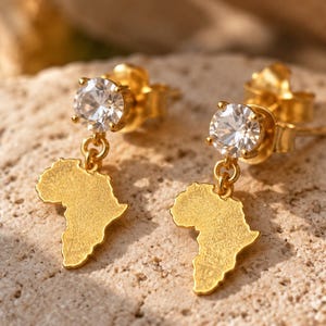 May include: A pair of gold-toned stud earrings featuring a round, clear, faceted gemstone at the top and a dangling, textured, flat charm shaped like the continent of Africa suspended from a small gold-toned jump ring.