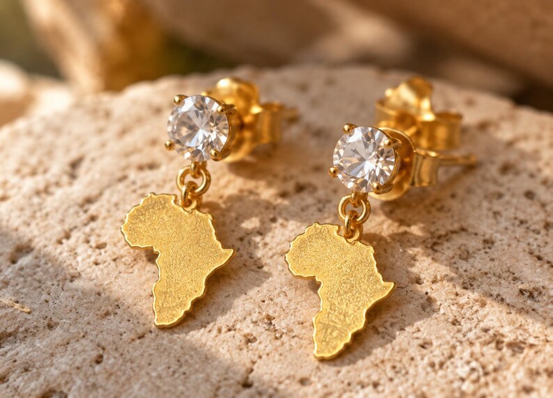May include: A pair of gold-toned stud earrings featuring a round, clear, faceted gemstone at the top and a dangling, textured, flat charm shaped like the continent of Africa suspended from a small gold-toned jump ring.