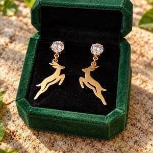 May include: A pair of gold-toned drop earrings featuring a leaping deer silhouette suspended from a round, clear, faceted cubic zirconia stud. The earrings are displayed inside a dark green velvet-lined jewelry box with a hinged lid.