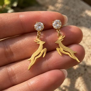May include: A pair of gold-toned drop earrings featuring a clear, round-cut gemstone stud at the top and a dangling, leaping deer charm suspended below. The deer charms have a textured, matte gold finish and are attached to the studs by small rings.