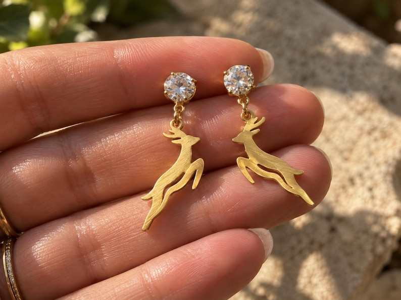 May include: A pair of gold-toned drop earrings featuring a clear, round-cut gemstone stud at the top and a dangling, leaping deer charm suspended below. The deer charms have a textured, matte gold finish and are attached to the studs by small rings.