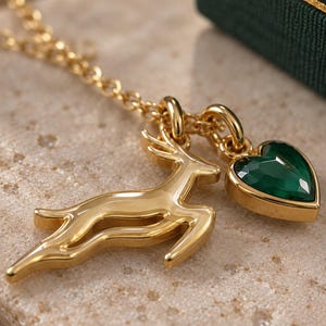 May include: A gold-toned necklace featuring two charms: a leaping deer silhouette and a faceted, heart-shaped dark green gemstone set in a gold-toned frame. The charms are suspended from a delicate gold-toned chain link necklace.
