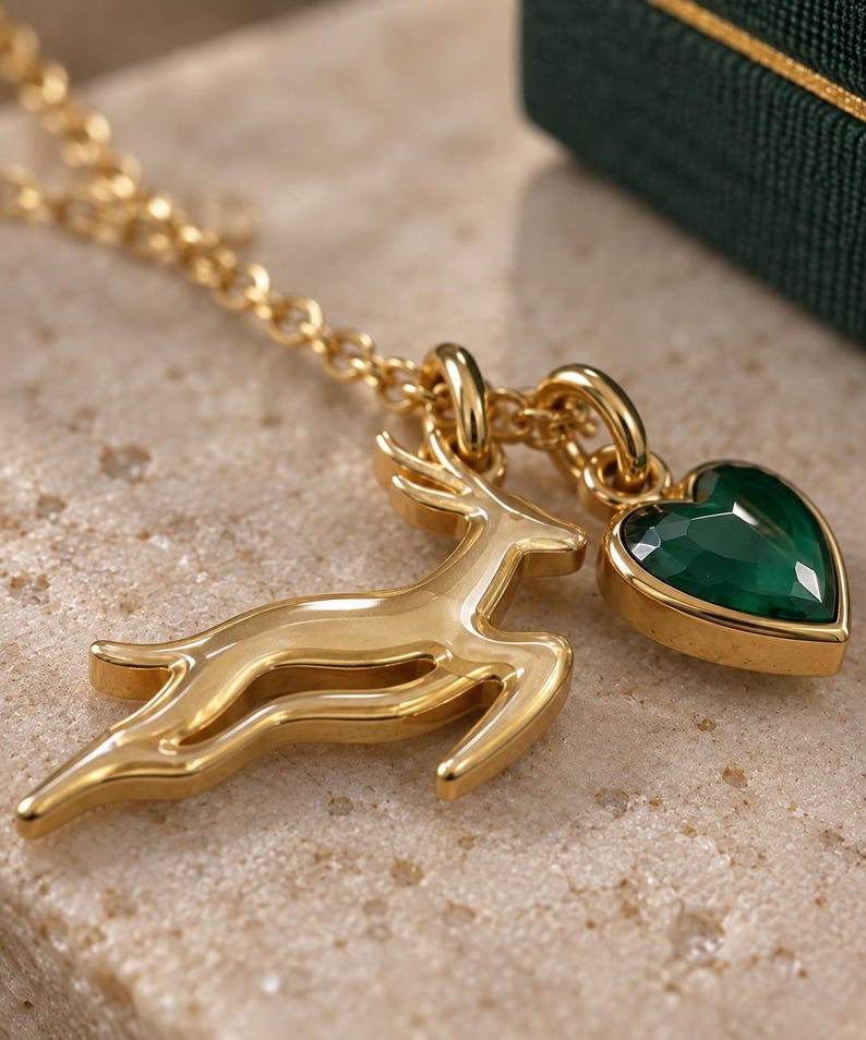 May include: A gold-toned necklace featuring two charms: a leaping deer silhouette and a faceted, heart-shaped dark green gemstone set in a gold-toned frame. The charms are suspended from a delicate gold-toned chain link necklace.