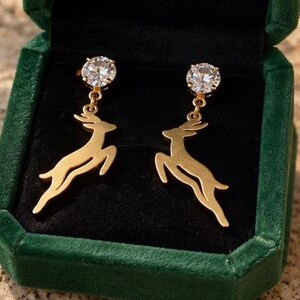 May include: A pair of gold-toned drop earrings featuring a leaping deer silhouette suspended from a round, clear, faceted stone stud. The earrings are displayed inside a dark green velvet-lined jewelry box with a black interior.