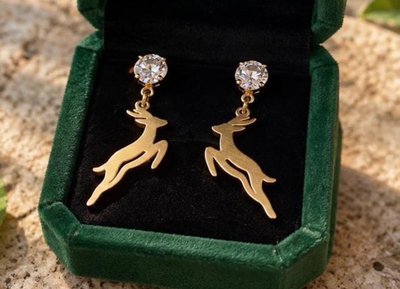 May include: A pair of gold-toned drop earrings featuring a leaping deer silhouette suspended from a round, clear, faceted stone stud. The earrings are displayed inside a dark green velvet-lined jewelry box with a black interior.