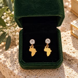 May include: A pair of gold-toned earrings featuring a textured map of Africa pendant suspended from a round, clear, faceted gemstone stud. The earrings are displayed inside a dark green velvet jewelry box with a black interior lining.