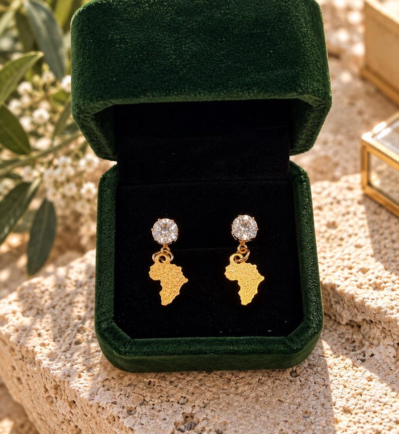 May include: A pair of gold-toned earrings featuring a textured map of Africa pendant suspended from a round, clear, faceted gemstone stud. The earrings are displayed inside a dark green velvet jewelry box with a black interior lining.