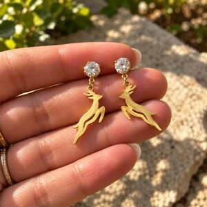 May include: A pair of gold-toned drop earrings featuring a leaping deer silhouette suspended from a single round, clear, faceted stone stud. The metal deer charms have a smooth, polished finish and are attached to the stone settings by small jump rings.
