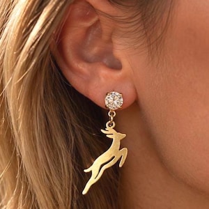 May include: A gold-toned earring featuring a round, clear, faceted stone stud connected to a dangling, flat, cutout charm in the shape of a leaping deer. The metal has a smooth, polished finish, and the design hangs vertically from the earlobe.
