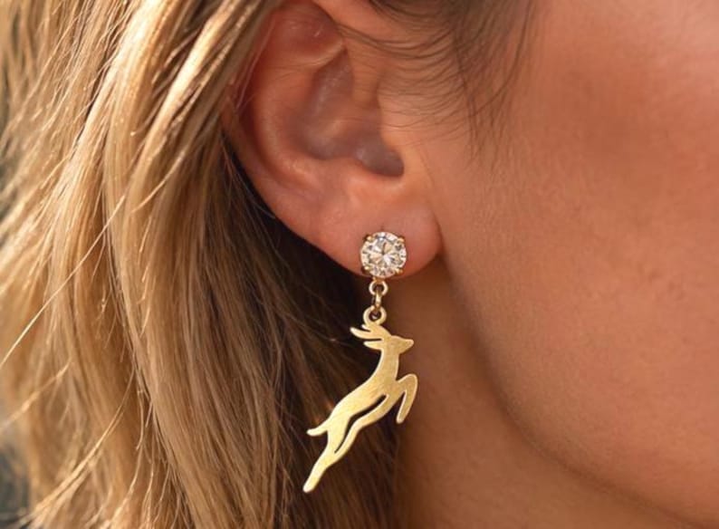 May include: A gold-toned earring featuring a round, clear, faceted stone stud connected to a dangling, flat, cutout charm in the shape of a leaping deer. The metal has a smooth, polished finish, and the design hangs vertically from the earlobe.
