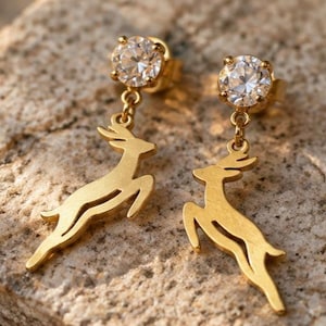 May include: A pair of gold-toned stud earrings featuring a round, clear faceted stone at the top. Dangling from each stone is a flat, gold-toned cutout charm in the shape of a leaping deer, suspended by a small metal link chain.