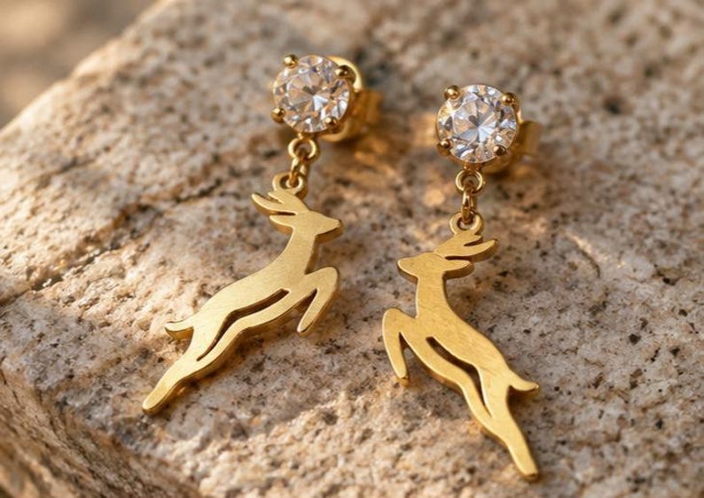 May include: A pair of gold-toned stud earrings featuring a round, clear faceted stone at the top. Dangling from each stone is a flat, gold-toned cutout charm in the shape of a leaping deer, suspended by a small metal link chain.