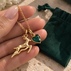 May include: A gold-toned necklace featuring two charms: a leaping deer silhouette and a faceted heart-shaped pendant in a deep emerald green color. The charms are suspended from a delicate gold-toned chain, creating a layered and detailed accessory.
