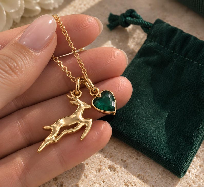 May include: A gold-toned necklace featuring two charms: a leaping deer silhouette and a faceted heart-shaped pendant in a deep emerald green color. The charms are suspended from a delicate gold-toned chain, creating a layered and detailed accessory.