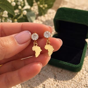 May include: A pair of gold-toned drop earrings featuring a round, clear, faceted stone stud attached to a dangling, flat, gold-toned charm shaped like the continent of Africa. The earrings are polished and have a reflective, metallic finish.