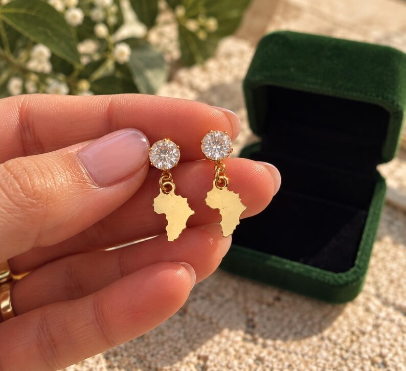 May include: A pair of gold-toned drop earrings featuring a round, clear, faceted stone stud attached to a dangling, flat, gold-toned charm shaped like the continent of Africa. The earrings are polished and have a reflective, metallic finish.