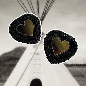 May include: Two heart-shaped earrings. Each earring features a black beaded border and a gold-colored heart in the center. The background is a black and white image of a teepee.
