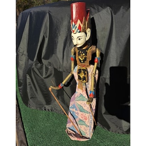 以下が含まれることがあります： 赤い高い帽子と塗装された顔を持つ木製の人形。人形は黒と金の装飾が施されたトップスと、ピンク、青、紫の色合いの模様のスカートを着用しています。2本の木の棒を持っています。