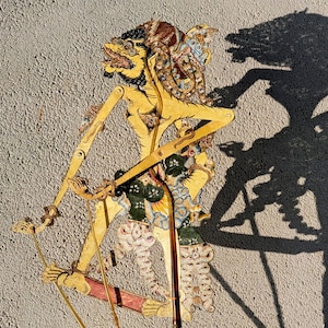 Op de afbeelding: Een traditionele Indonesische Wayang kulit-pop. De pop is gemaakt van ingewikkeld gesneden en beschilderd leer, met een gele body, gedetailleerde gezichtskenmerken en uitgebreide kostuumelementen. De pop wordt met staven vastgehouden.