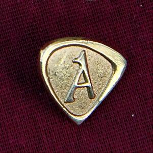 Può includere: Uno spillo in metallo dorato a forma di scudo con la lettera "A" al centro.