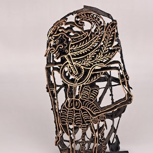 May include: An ornate black and gold metal batik stamp. The stamp features a stylized bird-like figure with intricate wing and body details. The design is set against a lattice background.