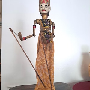 Puede incluir: Una marioneta de varilla Wayang Golek con rostro blanco y rasgos pintados, un tocado rojo alto y brazos de madera articulados. La marioneta viste un corpiño negro con lentejuelas florales y una falda larga de tela color mostaza con estampado.