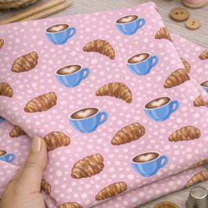 Puede incluir: Tela rosa con un patrón repetido de tazas de café azules con arte latte en forma de corazón y cruasanes marrones. La tela está salpicada de círculos blancos. Otros suministros de costura están en el fondo.
