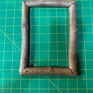 May include: A rectangular picture frame made from natural twigs. The frame is a muted brown color and has a rough, textured surface. The frame is set against a green grid background.