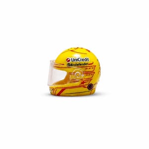 Pode incluir: Um capacete de corrida em miniatura amarelo com uma viseira transparente e detalhes gráficos vermelhos. O capacete exibe os logotipos da UniCredit e Bitdefender na lateral, juntamente com pequenos padrões de estrelas e linhas finas vermelhas.