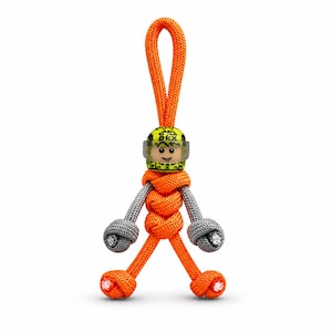 Puede incluir: Llavero hecho a mano naranja y gris con una cabeza de minifigura que lleva un casco amarillo con visera transparente. El llavero está hecho de cuerda anudada, con un lazo en la parte superior para colgar. La figura tiene brazos grises y piernas naranjas.