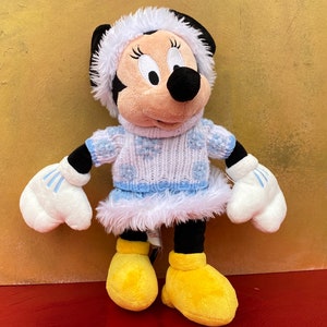 K&ouml;nnte beinhalten: Ein Pl&uuml;schtier von Mickey Mouse's Freundin Minnie Mouse, die einen wei&szlig;en und blauen Strickpullover mit Schneeflockenmuster, einen wei&szlig;en Pelzbesatz und einen wei&szlig;en Hut mit blauem Besatz tr&auml;gt.